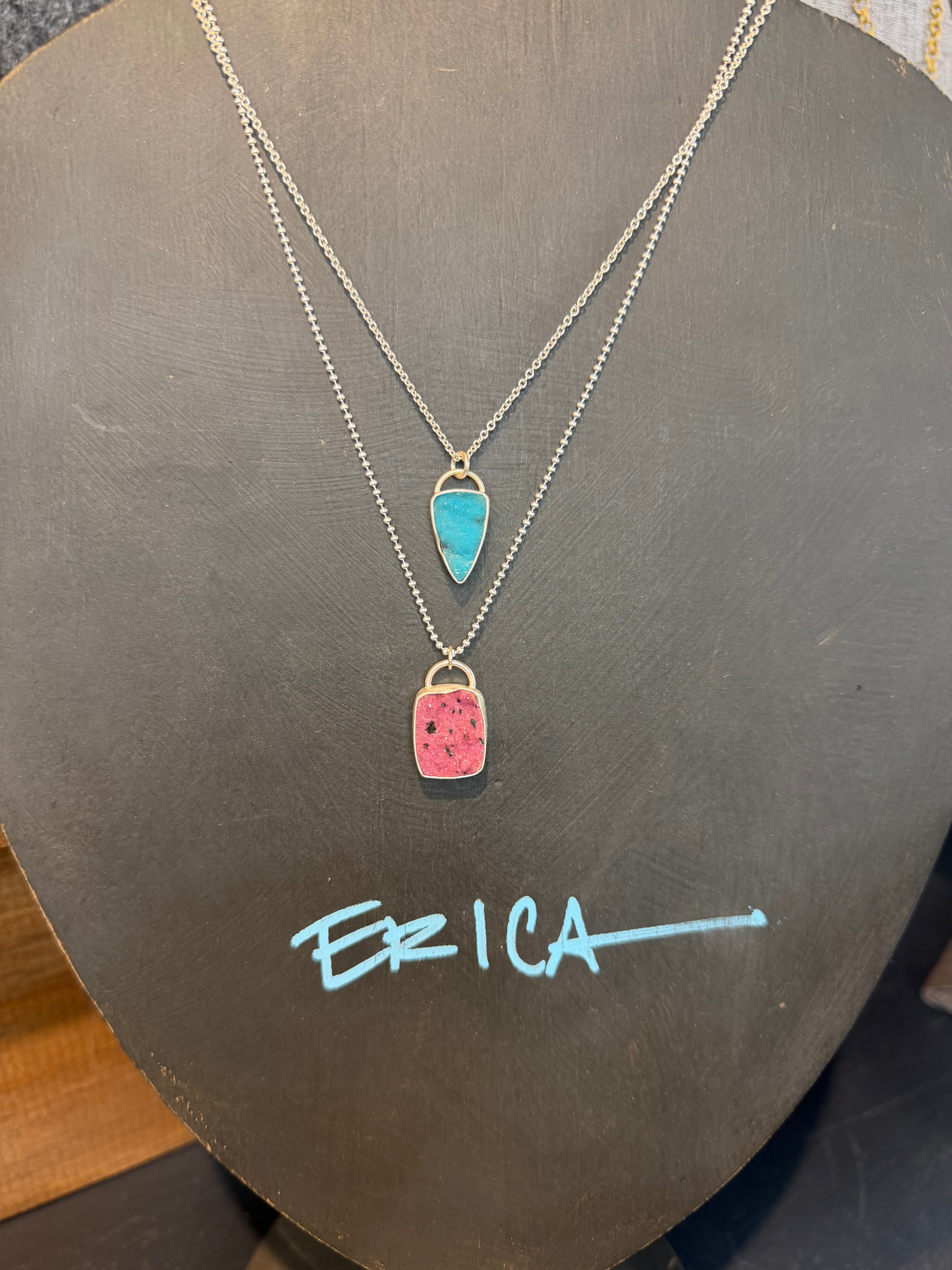 Two layered silver necklaces displayed on a dark gray jewelry bust. The top necklace features a small turquoise-blue teardrop pendant, and the bottom necklace features a larger pink square pendant with black speckles.