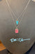 Two layered silver necklaces displayed on a dark gray jewelry bust. The top necklace features a small turquoise-blue teardrop pendant, and the bottom necklace features a larger pink square pendant with black speckles.